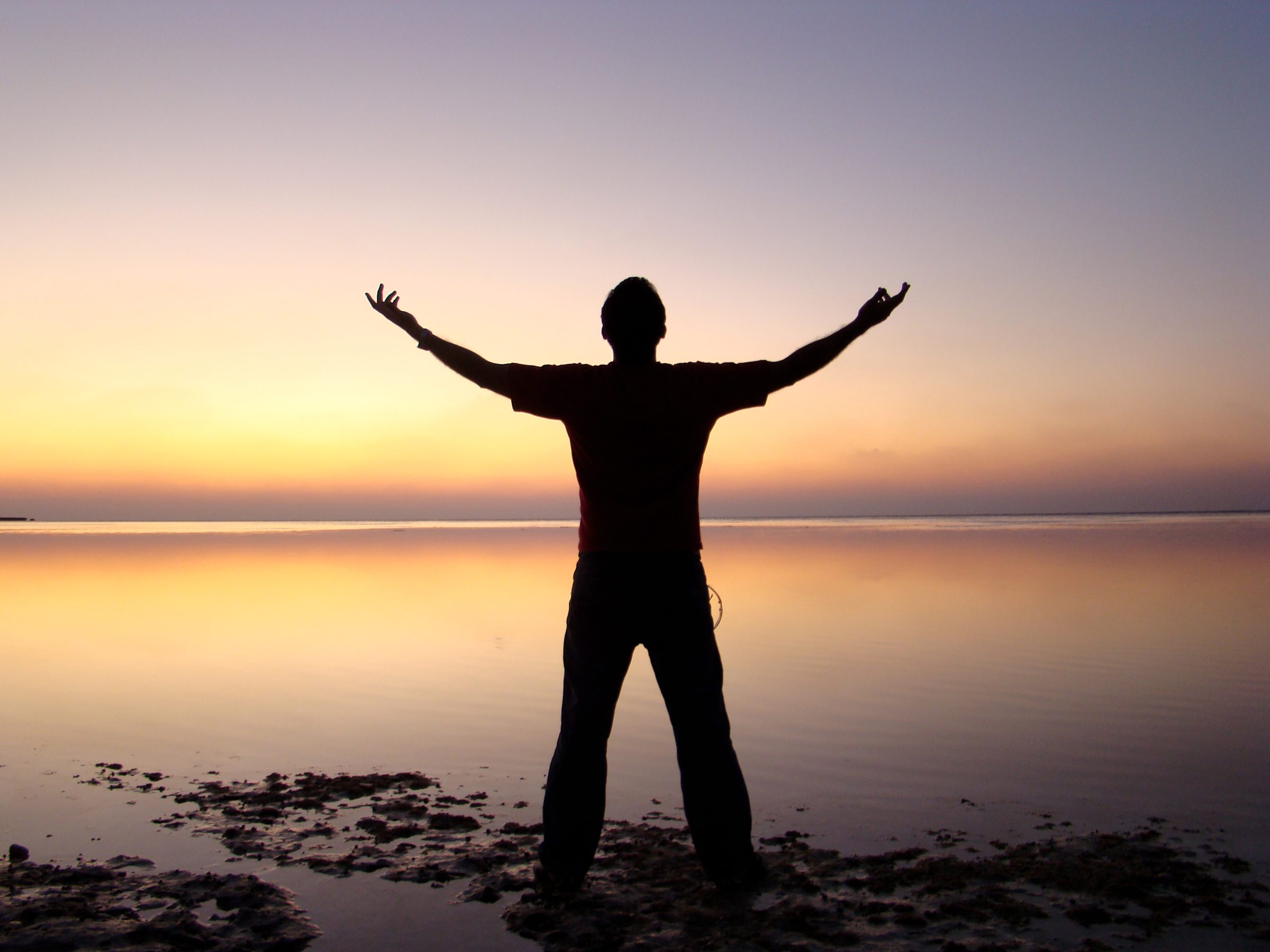 A man with arms raised in silhouette against a sunset over water